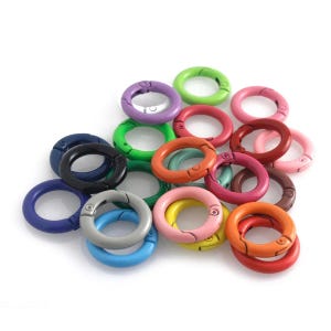 May include: A collection of colorful, round clasp rings in various shades including green, pink, orange, purple, and blue. These metal rings have a spring-loaded mechanism for easy opening and closing, ideal for crafting or attaching items.