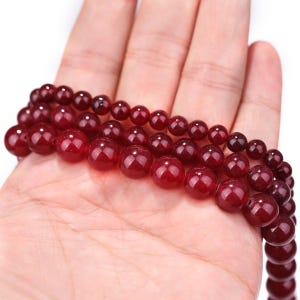 May include: Strands of polished, deep red gemstone beads. The beads are round and vary slightly in size, creating a textured appearance. The beads are held in a hand, showcasing their color and shine. Ideal for jewelry making.