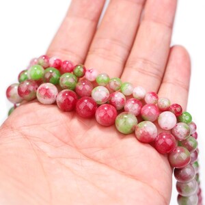 Puede incluir: Cadenas de cuentas de piedras preciosas en tonos rojos, verdes y blancos. Las cuentas son redondas y parecen pulidas. Las cuentas se sostienen en una mano, mostrando los colores y texturas de las piedras preciosas.