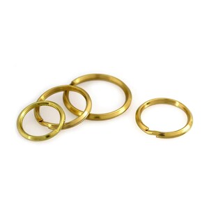 May include: A collection of gold-colored key rings of varying sizes, arranged on a white background. The key rings are circular, with one open for attaching keys. The metal has a shiny finish, reflecting light.