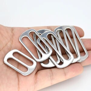 May include: A collection of shiny, silver-colored metal rectangle sliders. The sliders are held in a hand against a white background. The sliders have a rounded rectangular shape with a central opening.
