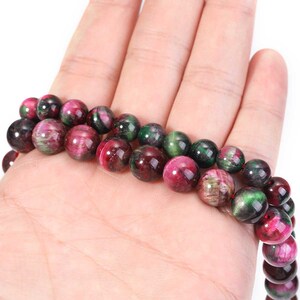 May include: Two strands of gemstone beads in shades of red, green, and black. The beads are round and appear to be polished. The beads are held in a hand, showcasing their color and texture. The beads are likely used for jewelry making.