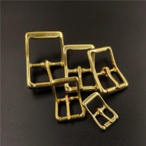 May include: A collection of shiny, gold-colored metal buckles in various sizes. The buckles have a rectangular frame with a central bar and a pin for securing straps. The hardware is displayed against a dark background.