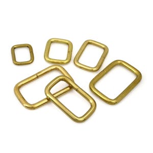 Puede incluir: Una colección de anillos metálicos rectangulares de color dorado de varios tamaños. Los anillos están dispuestos sobre una superficie blanca, con algunos superpuestos. El metal tiene un aspecto ligeramente envejecido, lo que sugiere un estilo vintage o antiguo. Estos anillos probablemente se utilizan para manualidades o ferretería.