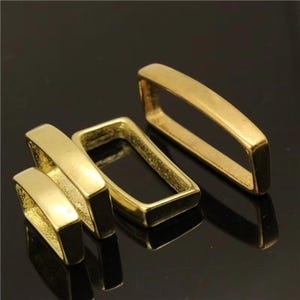 May include: Four gold-coloured rectangular metal buckles are arranged on a reflective black surface. The buckles have a smooth, polished finish and a slightly rounded shape. The lighting creates highlights and shadows.