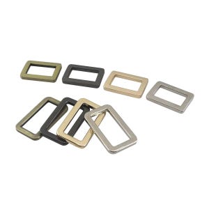 May include: A collection of rectangular metal buckles in various metallic finishes, including antique brass, black, gold, and silver. The buckles are arranged on a white background, showcasing their different colors and shapes. These are likely used for bags, belts, or other accessories.