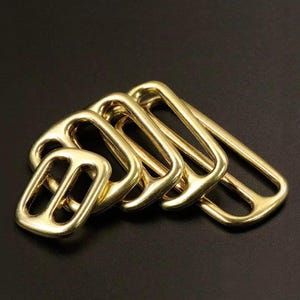 May include: A collection of five shiny, gold-colored metal buckles. Each buckle has a rectangular shape with a central bar and rounded edges. The buckles are arranged on a dark surface, with the light reflecting off their polished surfaces.