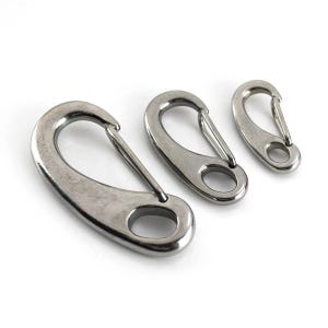 May include: Three stainless steel spring snap hooks of varying sizes are arranged on a white background. Each hook has a rounded body with a spring-loaded gate and a circular opening. The metal has a brushed finish.
