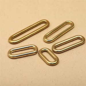 May include: Five gold-colored, oval-shaped metal rings. The rings are arranged on a light brown surface. The rings vary slightly in size, with a polished finish. These rings are likely used for crafting or jewelry making.