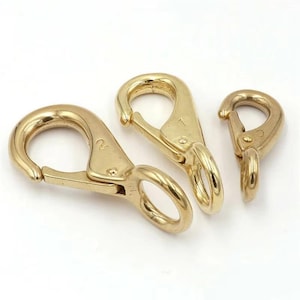 May include: Three brass spring snap hooks of varying sizes are displayed against a white background. The hooks have a shiny, golden finish and are designed with a spring-loaded mechanism for secure fastening. The largest hook is marked with the number "2".