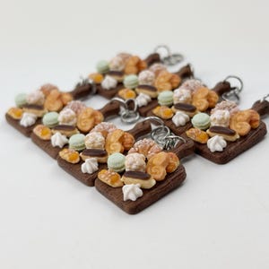 May include: A set of five miniature wooden cutting boards with a variety of miniature pastries, including croissants, eclairs, and macarons. The boards are brown and have a rustic look. Each board has a silver key ring attached.