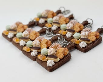 May include: A set of five miniature wooden cutting boards with a variety of miniature pastries, including croissants, eclairs, and macarons. The boards are brown and have a rustic look. Each board has a silver key ring attached.