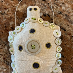 May include: A handmade, off-white fabric pincushion, adorned with numerous white buttons with green centers. The pincushion has a unique shape, with a loop at the top for hanging. Dark blue embroidered accents add contrast.