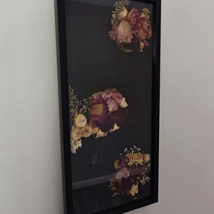 Puede incluir: Un marco rectangular negro tipo caja con tres arreglos separados de flores secas en tonos rojo oscuro, rosa apagado y crema. Las flores están dispuestas sobre un fondo oscuro detrás de un panel frontal de vidrio transparente.