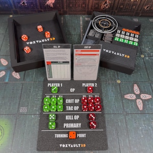 May include: A tabletop gaming setup with black trays, dice, and cards. The trays contain orange and white dice, and the cards display game information. Green and red dice are arranged on a game board with the text "VOXVAULT3D".