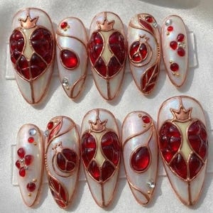 May include: A set of ten oval-shaped press-on nails. Each nail has a pearlescent white base and a pomegranate design. The design incorporates red, gold, and clear accents, with a crown detail on some. The nails are designed for easy application.