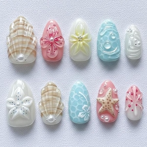 May include: A set of ten artificial nails with diverse designs. The nails showcase seashell, floral, and starfish decorations in pastel shades such as pink, blue, and white. Some nails have pearl accents.