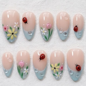 May include: A set of ten artificial nails with a light pink base, light blue tips, and various hand-painted designs. The designs include ladybirds, flowers, and water droplet accents. The nails are oval-shaped.