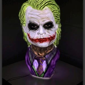 Puede incluir: Busto esculpido del Joker, con pelo verde, maquillaje blanco y una sonrisa roja. Viste un traje morado con corbata estampada. La pieza está iluminada desde dentro, creando un efecto brillante, un objeto decorativo.