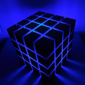 May include: A dark cube-shaped lamp with a grid pattern. The lamp emits a bright blue light from within the grid, creating a striking visual effect. The lamp is on a dark surface.