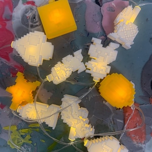 Può includere: Una stringa di luci LED pixelate bianche e gialle. Le luci hanno la forma di vari oggetti, tra cui quadrati e forme astratte. Le luci sono su un filo trasparente e sono disposte su una superficie colorata.