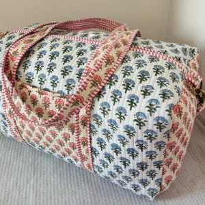 Può includere: Una borsa da viaggio trapuntata con un motivo floreale ripetuto di fiori blu e rosa su sfondo bianco. La borsa presenta manici in tessuto con bordo a righe rosa e bianche, una forma rettangolare e un attacco laterale in metallo dorato.