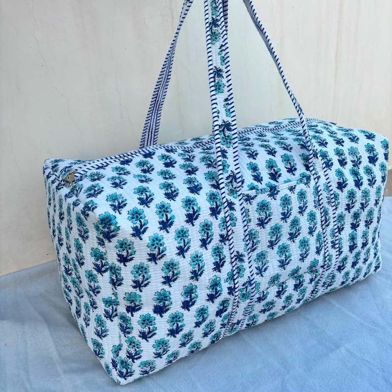 May include: A rectangular quilted duffel bag featuring a white base with a repeating floral pattern in shades of dark blue and bright turquoise. The bag includes two long fabric handles with a blue and white striped trim and a top zipper closure.