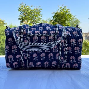 May include: A quilted duffel bag in a deep navy blue colour featuring a repeating pattern of light pink floral motifs. The bag includes two sturdy handles with a black and white striped trim that extends down the front of the quilted fabric.