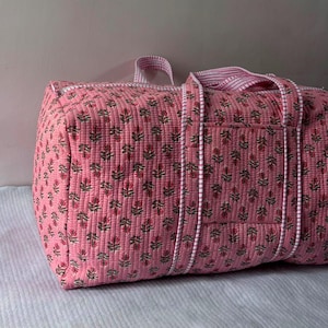 May include: A quilted duffel bag in a soft pink hue featuring a repeating floral pattern in shades of green and dark pink. The bag includes white and pink striped fabric handles and trim, with a rectangular exterior pocket on the side panel.