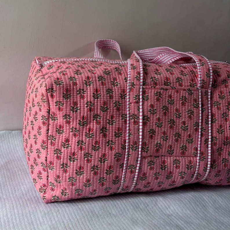 May include: A quilted duffel bag in a soft pink hue featuring a repeating floral pattern in shades of green and dark pink. The bag includes white and pink striped fabric handles and trim, with a rectangular exterior pocket on the side panel.