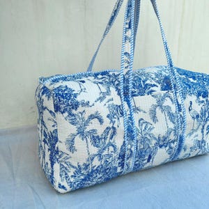May include: A rectangular quilted duffel bag featuring a white base with a blue toile-style print depicting tropical palm trees, birds, and tigers. The bag includes two long shoulder straps with a blue and white striped pattern along the edges.
