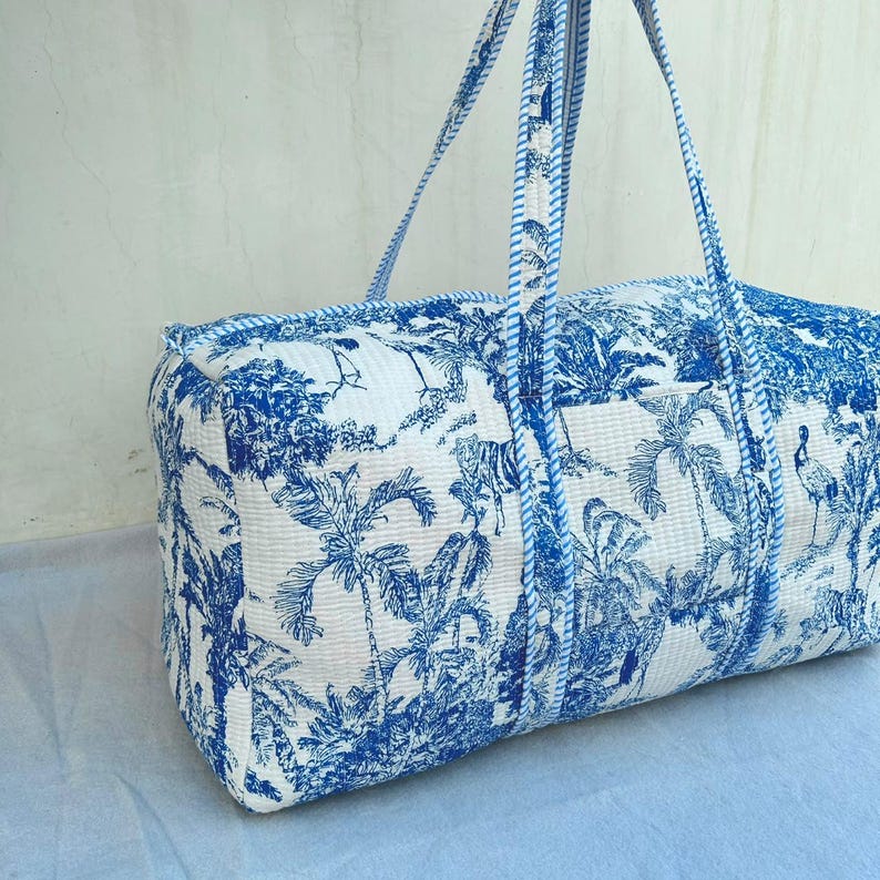 May include: A rectangular quilted duffel bag featuring a white base with a blue toile-style print depicting tropical palm trees, birds, and tigers. The bag includes two long shoulder straps with a blue and white striped pattern along the edges.