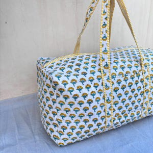 May include: A rectangular quilted duffel bag featuring a white base with a repeating pattern of small yellow and teal floral motifs. The bag includes two long fabric handles with a yellow and white striped trim that matches the bag's edges.