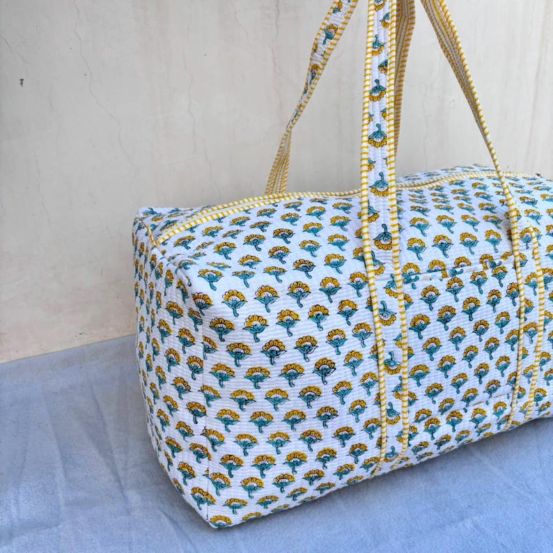 May include: A rectangular quilted duffel bag featuring a white base with a repeating pattern of small yellow and teal floral motifs. The bag includes two long fabric handles with a yellow and white striped trim that matches the bag's edges.