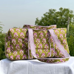 May include: An olive green quilted tote bag featuring a floral design in purple and white. The bag has a rectangular shape, a top handle, and a shoulder strap. The straps are white with purple stripes.
