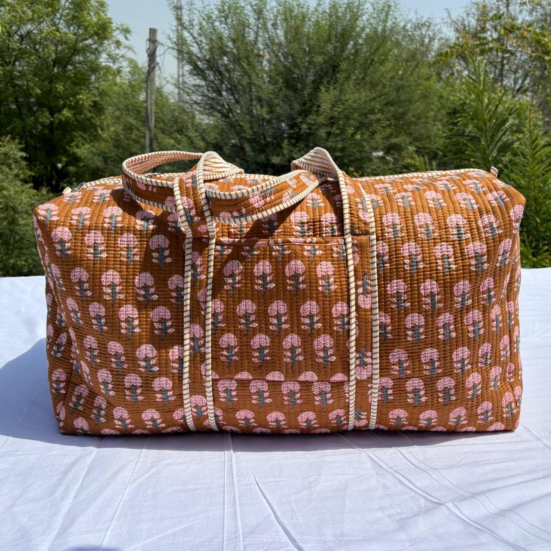 May include: A rectangular quilted duffel bag in a warm brown tone featuring a repeating pattern of small pink and white floral motifs. The bag includes two sturdy fabric handles and a large exterior pocket on the front with striped trim.