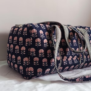 May include: A quilted duffel bag in a dark navy blue fabric featuring a repeating pattern of light pink floral motifs. The bag includes two sturdy handles with a black and white striped trim and a matching zipper closure along the top edge.