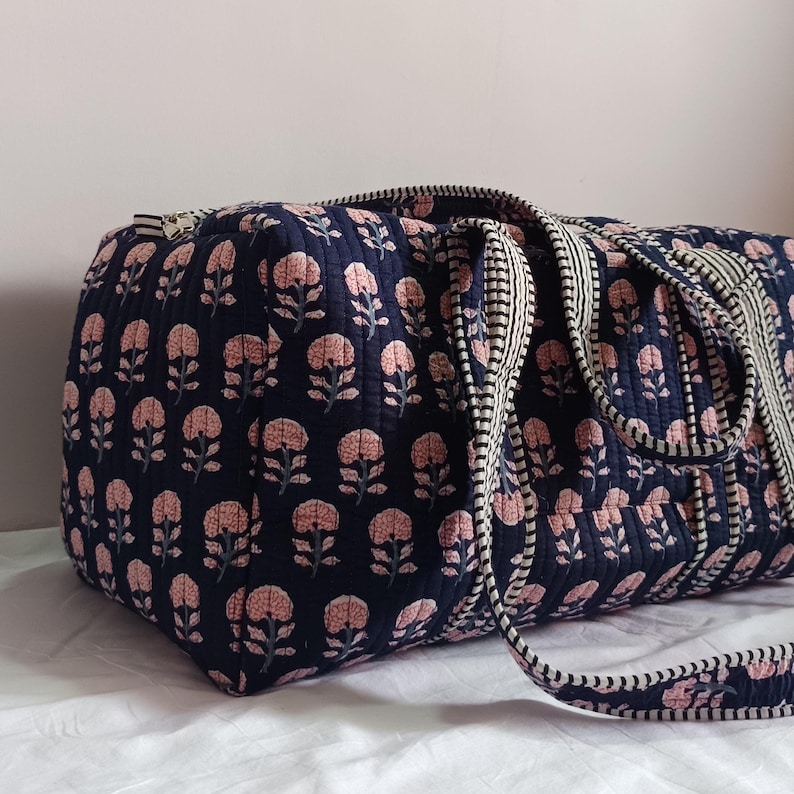 May include: A quilted duffel bag in a dark navy blue fabric featuring a repeating pattern of light pink floral motifs. The bag includes two sturdy handles with a black and white striped trim and a matching zipper closure along the top edge.