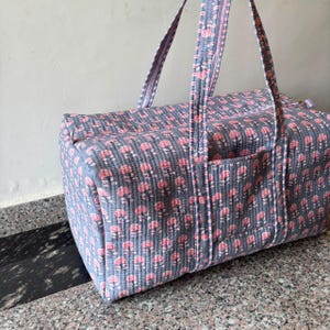 May include: A rectangular quilted duffel bag featuring a repeating pattern of small pink floral motifs on a slate blue background. The bag includes two long shoulder straps with a striped trim and a zippered top closure with a side pocket.