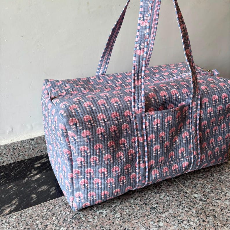 May include: A rectangular quilted duffel bag featuring a repeating pattern of small pink floral motifs on a slate blue background. The bag includes two long shoulder straps with a striped trim and a zippered top closure with a side pocket.