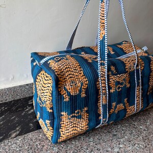 May include: A quilted duffel bag featuring a repeating pattern of orange and black tigers on a dark blue background with vertical stripes. The bag includes two long carrying handles made of white and blue striped fabric and a top zipper closure.