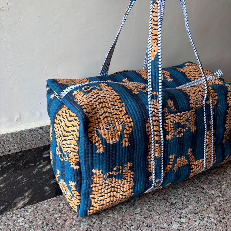 May include: A quilted duffel bag featuring a repeating pattern of orange and black tigers on a dark blue background with vertical stripes. The bag includes two long carrying handles made of white and blue striped fabric and a top zipper closure.