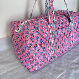 May include: A quilted duffel bag featuring a vibrant pink background with a repeating pattern of blue floral motifs. The bag includes a top zipper closure and two long carrying straps with a black and white striped design along the edges.