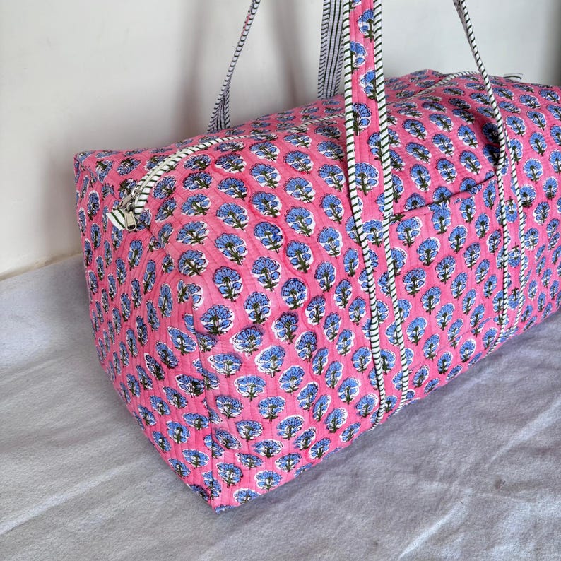 May include: A quilted duffel bag featuring a vibrant pink background with a repeating pattern of blue floral motifs. The bag includes a top zipper closure and two long carrying straps with a black and white striped design along the edges.