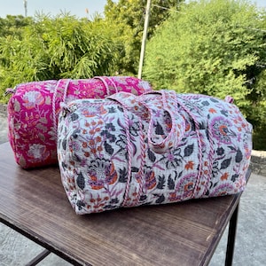 Könnte beinhalten: Zwei Reisetaschen mit Blumenmuster. Eine Tasche ist leuchtend rosa mit einem sich wiederholenden Blumenmuster. Die andere Tasche ist weiß mit einem Blumenmuster in Lila-, Orange- und Schwarztönen. Beide Taschen haben passende rosa Seilgriffe.