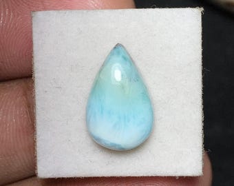 May include: A teardrop-shaped, polished gemstone with a gradient of colors, from light blue to white. The stone is set on a square, off-white surface, showcasing its smooth, reflective surface. The gemstone is likely a pendant or a jewelry component.