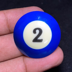 May include: A blue billiard ball with a white circle featuring the number 2 in black. The ball is a solid blue color with a glossy finish. This pool ball is a standard size for playing pool.