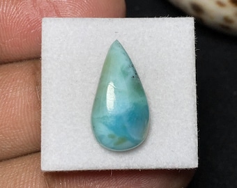 May include: A teardrop-shaped gemstone with a polished surface. The stone features a blend of light blue and turquoise hues, with darker blue swirls and subtle green accents. It is set on a white square.