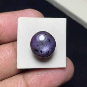 May include: A round, cabochon-cut purple gemstone with a smooth, polished surface. The stone features a distinct star-like light reflection and visible dark, needle-like inclusions throughout its translucent, deep violet body.