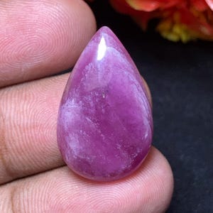 Könnte beinhalten: Ein polierter, tropfenförmiger Edelstein in tiefen Rosa- und Lavendeltönen. Der Stein hat eine glatte, glänzende Oberfläche mit innerer Marmorierung. Der Edelstein wird zwischen den Fingern gehalten, um seine Größe und Farbe zu zeigen.